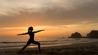 Woman practicing warrior yoga pose on beach at sunset with dramatic orange sky and ocean waves creating peaceful meditation scene.