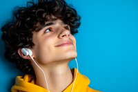 Young Hispanic male with curly dark hair wearing yellow hoodie listening to music through white earphones against bright blue background, looking up with joyful expression.