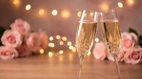 Two champagne flutes filled with sparkling wine against romantic pink roses and warm bokeh lights creating elegant celebration atmosphere for weddings.