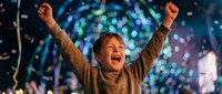 Young boy celebrating with raised arms under colorful disco lights at night party event. Joyful child expressing excitement and happiness during festive celebration.