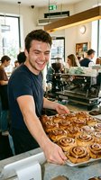 Young male barista of Caucasian ethnicity displaying freshly baked cinnamon rolls at counter in busy cafe. Professional bakery worker smiling while presenting pastries in modern coffee shop with customers and espresso machine.