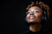 Young African American woman with natural dreadlocks smiling while listening to music through headphones against dark background, expressing joy and contentment.