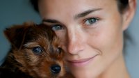 Young Caucasian woman with natural makeup and gentle smile poses cheek to cheek with adorable brown Yorkshire terrier puppy with bright blue eyes in intimate close-up portrait.