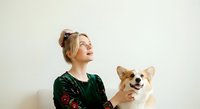 Woman with blonde hair in a bun, Caucasian ethnicity, holding and posing with a tan and white Corgi dog against a neutral white background for pet adoption or lifestyle content.