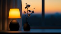 Cozy evening atmosphere with glowing table lamp and silhouette of dried flowers in vase against sunset window view, creating warm minimalist home decor setting.
