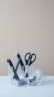 Clear acrylic desk organizer with office supplies including pens, scissors, tape dispenser and paper clips on white surface against beige background.
