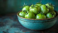 Fresh green unripe tomatoes in rustic turquoise ceramic bowl against teal background, perfect for farm-to-table food concepts.