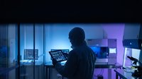 Male cybersecurity analyst monitoring network security systems on multiple screens in modern tech office with blue ambient lighting at night.