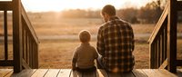 Father and son sitting together on wooden deck during golden hour sunset, bonding moment in rural countryside setting with warm lighting.