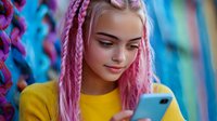 Young woman with pink braided hair wearing yellow sweater looking at smartphone against colorful blurred background, modern urban lifestyle portrait.