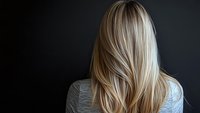 Back view of blonde woman with long straight hair against dark background, showcasing healthy hair with highlights and lowlights for salon advertising.
