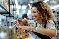 Young mixed race female bartender with curly hair smiling while mixing drinks at modern bar counter, wearing apron and casual attire in professional setting.