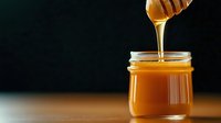 Golden honey dripping from wooden dipper into glass jar on dark background, natural sweetener for healthy cooking and organic food preparation.