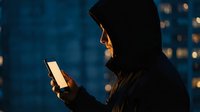 Hooded person using smartphone at night in urban setting with city lights creating atmospheric bokeh background for technology and privacy concepts.