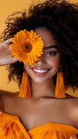 Young African American woman with natural curly hair holding orange gerbera daisy near eye, wearing matching orange dress and tassel earrings against yellow background, bright smile.