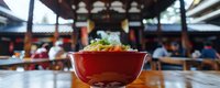 Traditional red bowl with fresh salad in Asian restaurant setting, blurred patrons dining in ornate temple-like structure with wooden beams and cultural architecture.