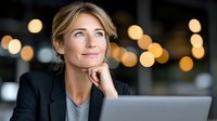 Professional blonde businesswoman looking up thoughtfully while working on laptop in modern office with warm bokeh lights in background.