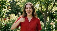 Happy young Caucasian woman in red shirt giving thumbs up gesture outdoors in green garden setting with natural sunlight for positive lifestyle content.