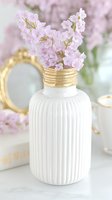 Delicate pink flowers in white ribbed ceramic vase with gold neck detail, styled with vintage mirror and soft pastel background for elegant home decor and lifestyle photography.