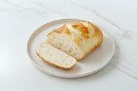 Fresh crusty white bread loaf sliced on round ceramic plate against white marble background, showing soft interior texture and golden crust surface.
