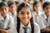 Young South Asian student in school uniform with braided hair and bright smile, sitting in classroom among classmates. Natural lighting highlights genuine expression.