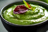 Creamy green avocado sauce in black ceramic bowl garnished with fresh herbs and red leaf. Close-up macro shot with selective focus on smooth texture.