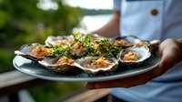 Fresh oysters on half shell garnished with herbs and sauce served on plate by chef in white uniform against blurred outdoor background.