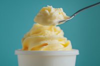 Creamy soft serve vanilla ice cream swirled in white cup with spoon scooping from top, captured against vibrant turquoise background. Perfect for dessert advertising.
