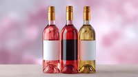 Three wine bottles in rose, red and white varieties with blank labels arranged on light surface against soft pink bokeh background. Suitable for wine marketing.