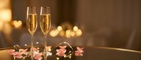 Two elegant champagne flutes filled with sparkling wine on dark surface with pink rose petals and warm golden bokeh lights creating romantic atmosphere.