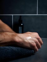 Wet male hand resting on dark towel in spa setting with massage oil bottle in background, creating atmospheric wellness and relaxation scene against black tile wall.