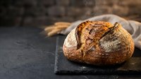 Freshly baked artisan sourdough bread loaf on dark slate board with wheat ears and linen cloth creating rustic bakery atmosphere for food photography.