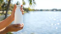 Person holding white reusable water bottle near tropical waterway with palm trees and sparkling water, promoting eco-friendly hydration and sustainable lifestyle choices outdoors.