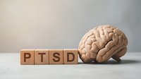 Wooden blocks spelling PTSD next to a human brain model on a gray surface, symbolizing post-traumatic stress disorder awareness and mental health.