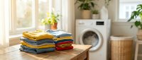 Stack of neatly folded colorful clothes on wooden table in bright laundry room with washing machine and plants. Household chores and domestic life concept.