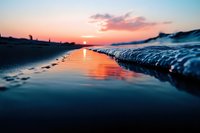Dramatic coastal sunset reflection on wet sand beach with gentle wave creating mirror effect, orange and blue sky colors merge with dark silhouetted shoreline.