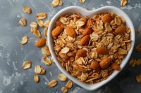 Homemade granola with almonds and oat flakes in heart shaped ceramic bowl on dark background. Healthy breakfast concept with scattered ingredients.
