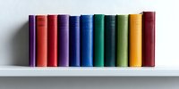 Row of colorful hardcover books standing in line on white shelf against neutral wall background, arranged in rainbow spectrum order showing spines with minimal design.