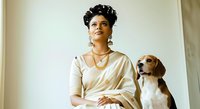 Young woman with dark curly hair and South Asian ethnicity wearing cream-colored draped clothing, posing with a beagle dog against neutral background for lifestyle and pet companionship concepts.