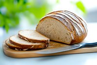 Fresh homemade sourdough bread loaf sliced on wooden cutting board with knife against bright kitchen background, selective focus on crusty surface.