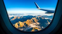 Aerial view from airplane window showing snow-capped mountain peaks bathed in golden sunlight with wing visible, suitable for travel and aviation content.