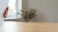 Woman's hand peeking around white wall corner in modern interior space with wooden floor and blurred green plant background.