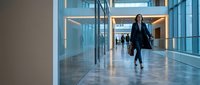 Professional businesswoman walking through modern office corridor with glass walls and contemporary architecture for corporate presentations.