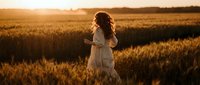 Young woman with curly hair in white dress standing in golden wheat field during sunset with mountains in background creating warm atmospheric scene.