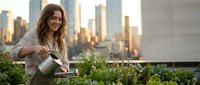Young woman watering plants on urban rooftop garden with city skyline at sunset. Professional gardening and sustainable living concept for eco-friendly lifestyle.