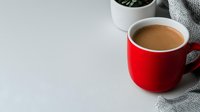 Red ceramic mug filled with hot coffee or cocoa drink on light background with gray knitted textile and green plant, copy space for text.