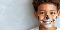 Young boy with curly hair wearing creative skeleton face paint design smiling against light gray wall background. Halloween makeup art for children.