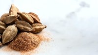 Whole cardamom pods and ground cardamom spice powder scattered on white marble surface, macro food photography showing texture and detail of aromatic spices.