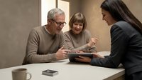 Senior man with gray hair and glasses wearing tan sweater, middle-aged woman with brown hair wearing beige cardigan, and young woman with long dark hair wearing dark blazer reviewing documents together at table for business meeting or consultation.