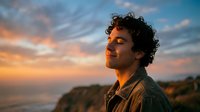 Young Hispanic man with curly hair enjoying peaceful sunset moment on coastal cliff, eyes closed in meditation and mindfulness practice outdoors.